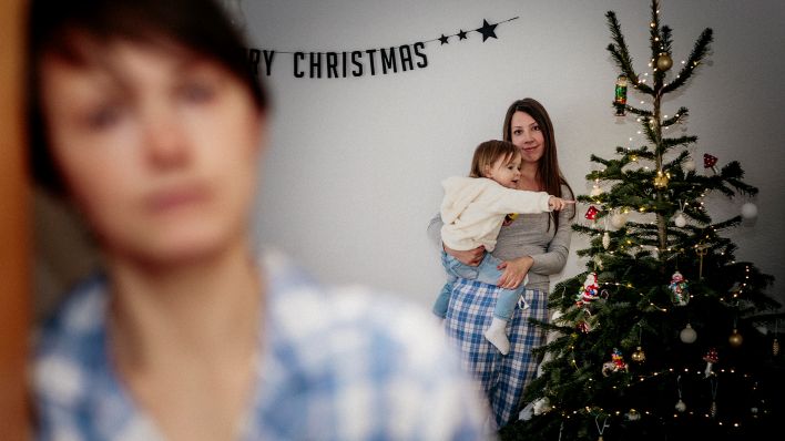 Archivbild: Eine Frau steht, unscharf zu sehen, im Vordergrund; ihre Tochter steht, mit Enkeltochter auf dem Arm, im Hintergrund, neben einem Weihnachtsbaum. (Quelle: dpa/Mikhrin)
