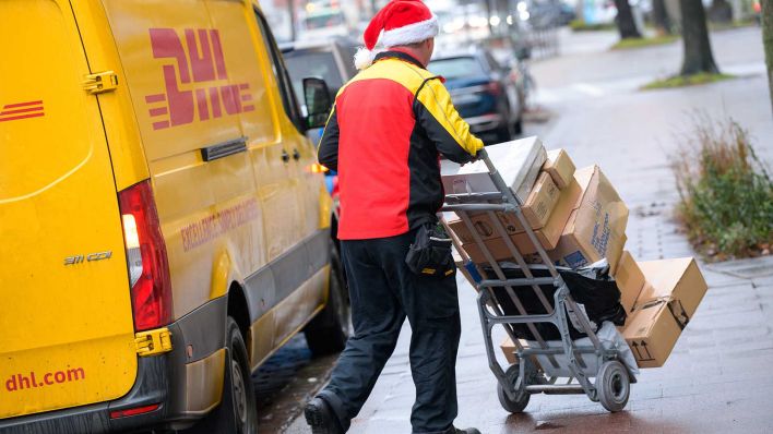 Archivbild: Ein Paketbote des Paketdienstes DHL schiebt mit einer Weihnachtsmütze auf dem Kopf seine Fracht an seinem Transporter vorbei. (Quelle: dpa/Walzberg)
