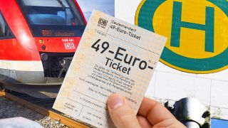 Symbolbild: Hand hält 49-Euro-Ticket vor Regionalbahn und Bushaltestellenschild. (Quelle: dpa/Ohde)