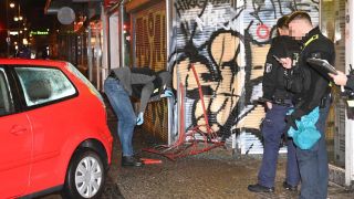 Der Tatort: Links ist das Tatauto zu sehen, in der Mitte ein Polizeibeamter, der den Fahrradständer und den Rollladen des Geschäfts inspiziert. Bild: Morris Pudwell