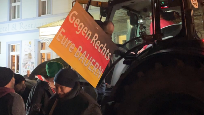 Es sind Demonstranten auf einer Demo zu sehen, darunter auch Bauern. Es ist ein Traktor im Hintergrund zu erkennen und es wird ein Schild hochgehalten, auf dem steht " Gegen rechts eure Bauern".
