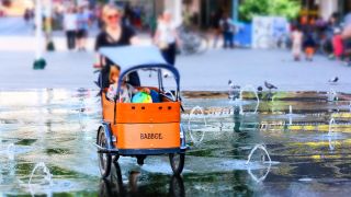 Symbolbild:Eine Frau fährt ihr Kind mit einem Babboe-Lastenrad durch einen Wasserbrunnen.(Quelle:imago images/M.Gstettenbauer)