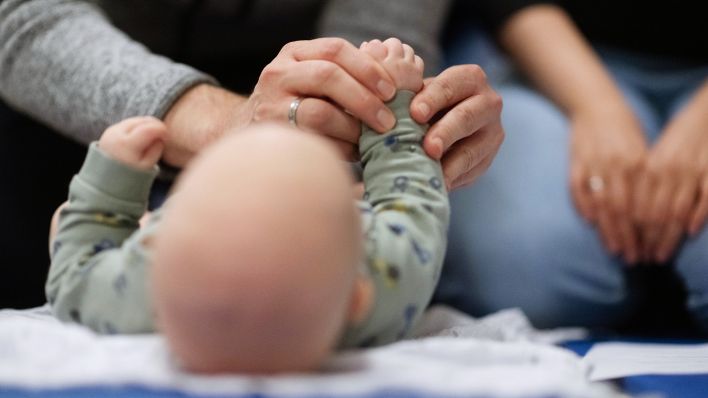 Symbolbild:Ein Vater hält während een Infant Handling Curses die Füße seines Kindes, während die Mutter daneben sitzt.(Quelle:picture Alliance/dpa/M.Murat)