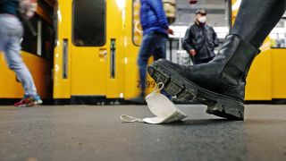 Symbolbild:Auf einem Bahnsteig vor einer haltenden U-Bahn tritt ein Stiefel auf eine am Boden liegende FFP2-Maske.(Quelle:imago images/S.Gudath)