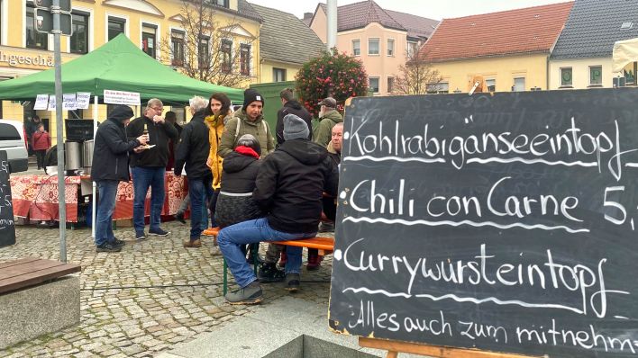 Besucher der Suppenmeile sitzen an Tischen oder stehen und essen, im Vordergrund ein Schild mit angebotenen Suppen (Foto: rbb/Schilka)