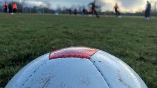 Ein Fußball liegt im vorderen Bildrand, dahinter sieht man verschwommen Menschen auf einem Rasen trainieren.