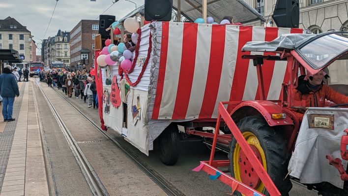 Ein bunt geschmückter Karnevalswagen mit dem Cottbuser Rathaus im Hintergrund wird von einer Gruppe verkleideter Kinder gefolgt.